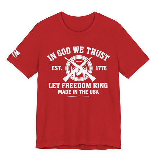 Red unisex t-shirt with white “IN GOD WE TRUST” crossed-rifles graphic and “LET FREEDOM RING” text, left-sleeve flag graphic, white background.