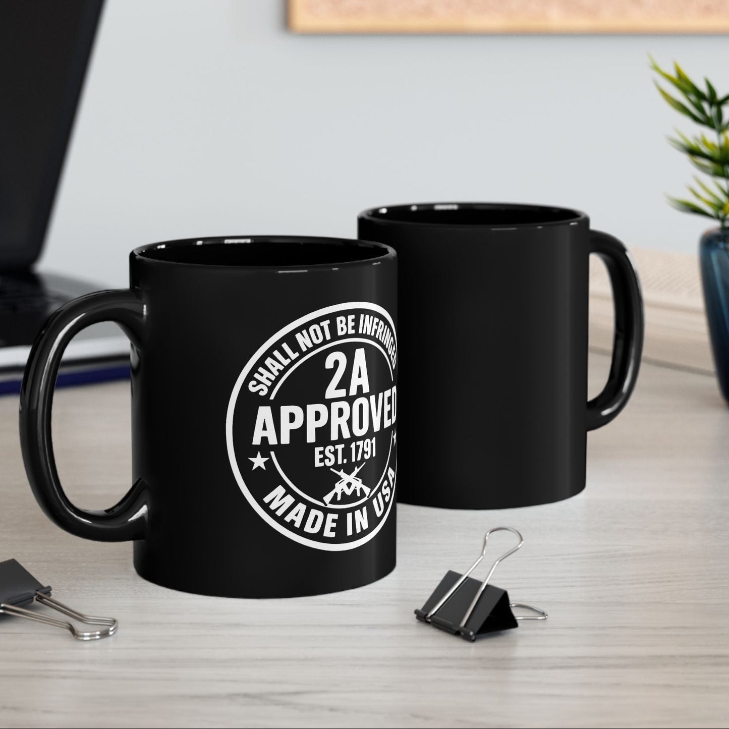 Two black ceramic mugs on an office desk; front mug shows “2A Approved” white design