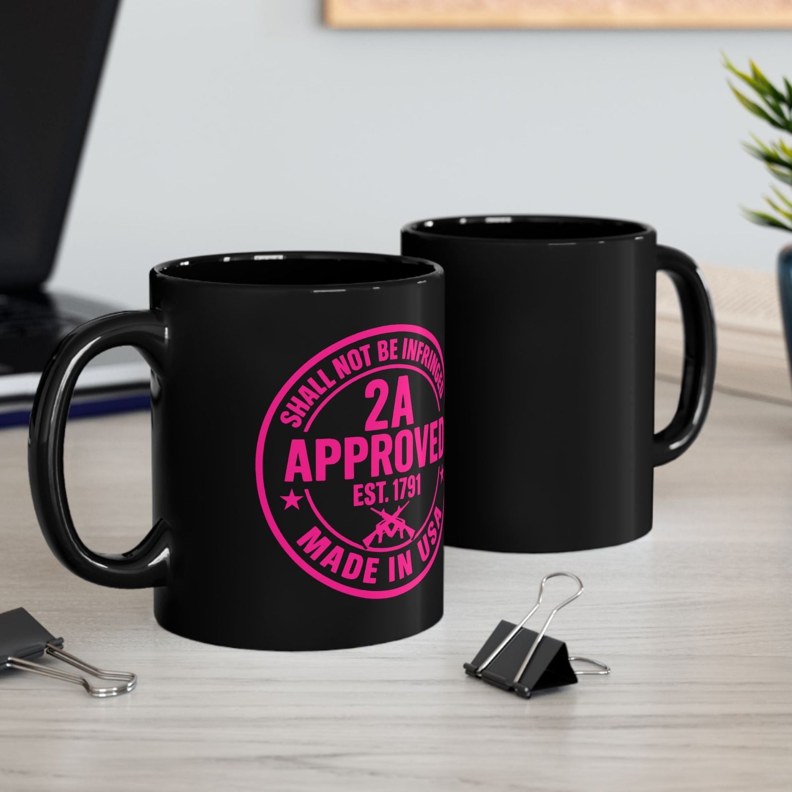 Two black ceramic mugs on an office desk; front mug shows pink “2A Approved” design