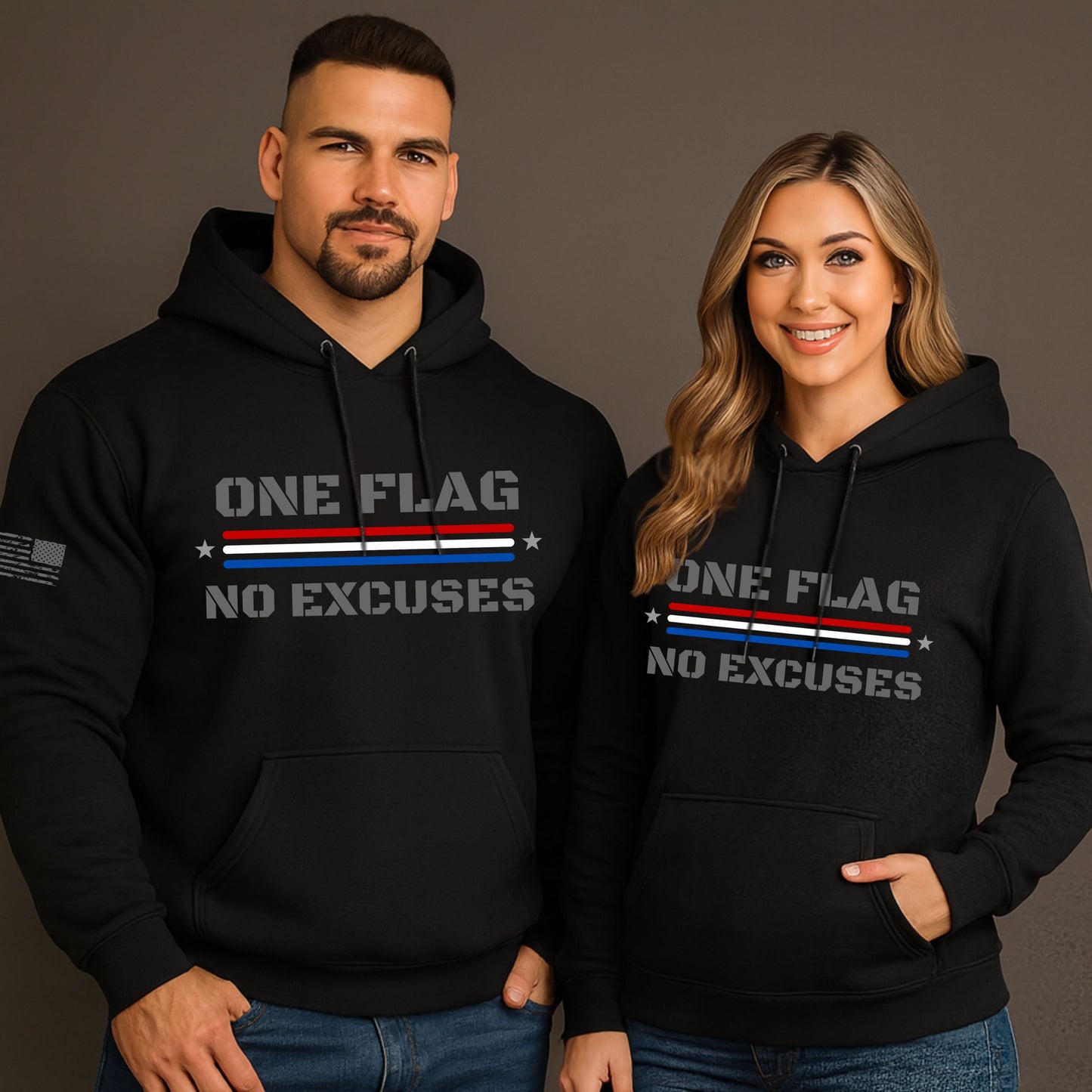 Man and woman wearing matching black ‘One Flag No Excuses’ hoodies with subdued American flag design, standing in front of a warm gray studio background.